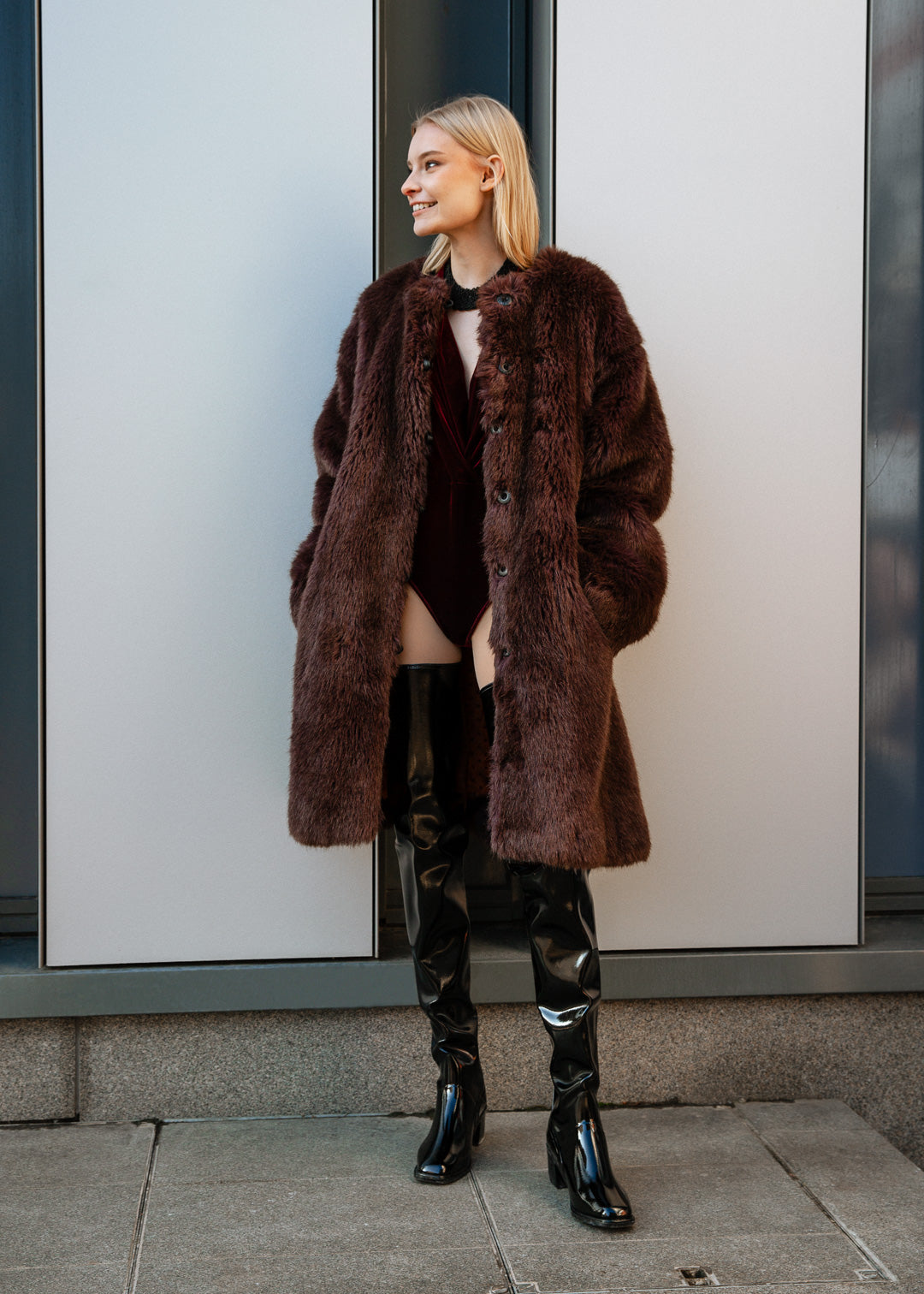 Model wearing ACQUO of Sweden over-the-knee glowing black heeled boots, styled with a deep red velvet bodysuit and oversized brown faux fur coat – a bold, luxurious street style look with Scandinavian elegance