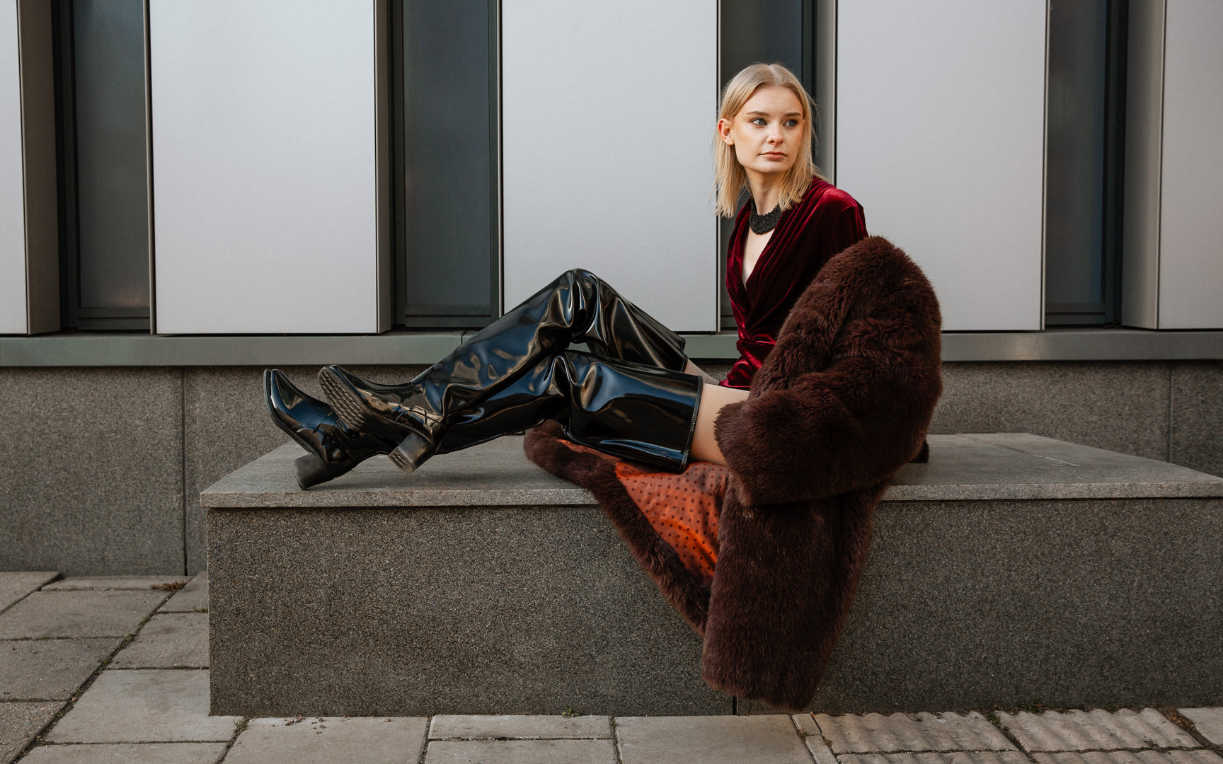 Model wearing ACQUO of Sweden over-the-knee black heeled boots, styled with a deep red velvet bodysuit and oversized brown faux fur coat – a luxurious and modern fashion statement captured in an urban setting