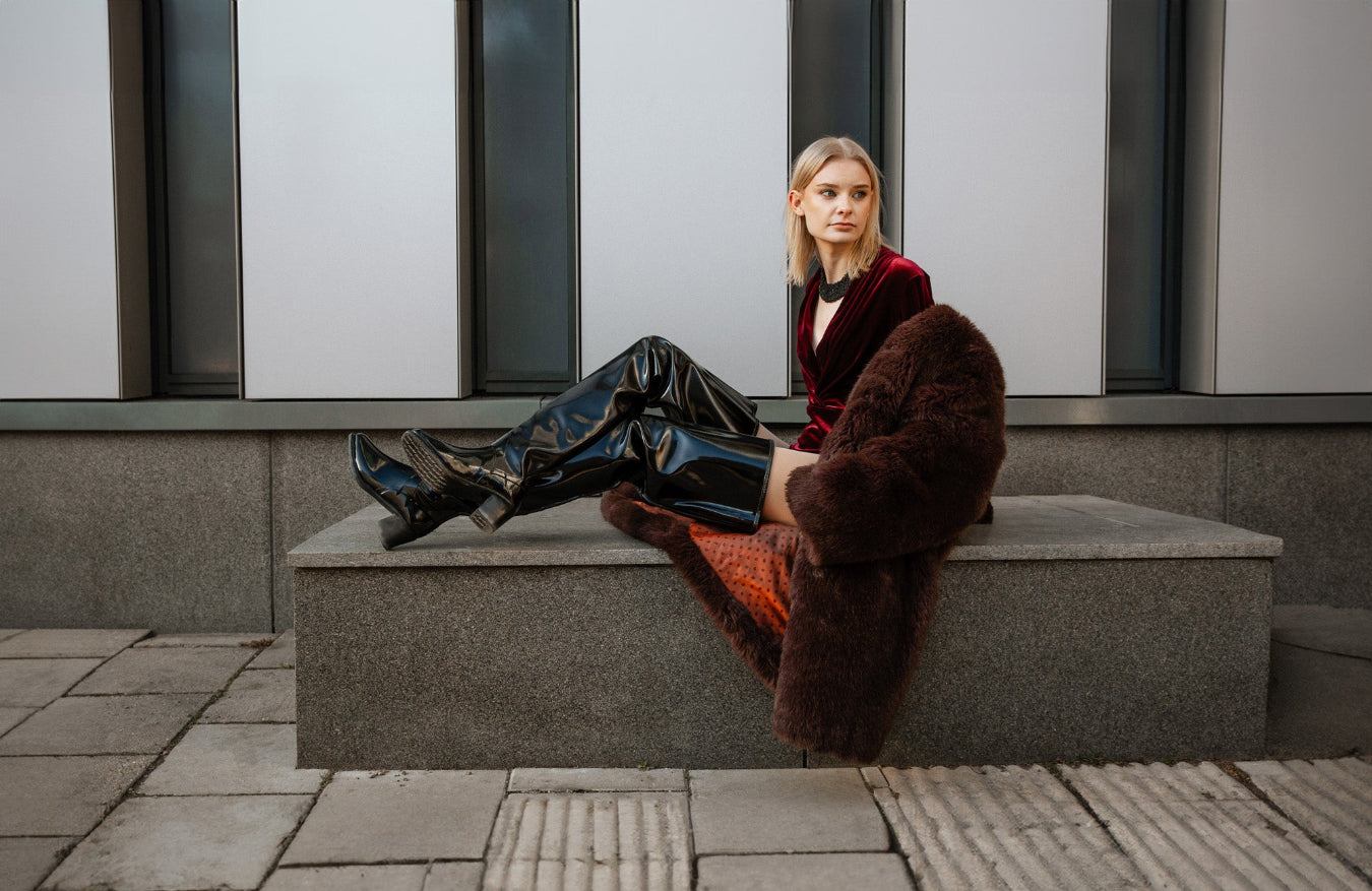 Model wearing ACQUO of Sweden over-the-knee black heeled boots, styled with a deep red velvet bodysuit and oversized brown faux fur coat – a luxurious and modern fashion statement captured in an urban setting