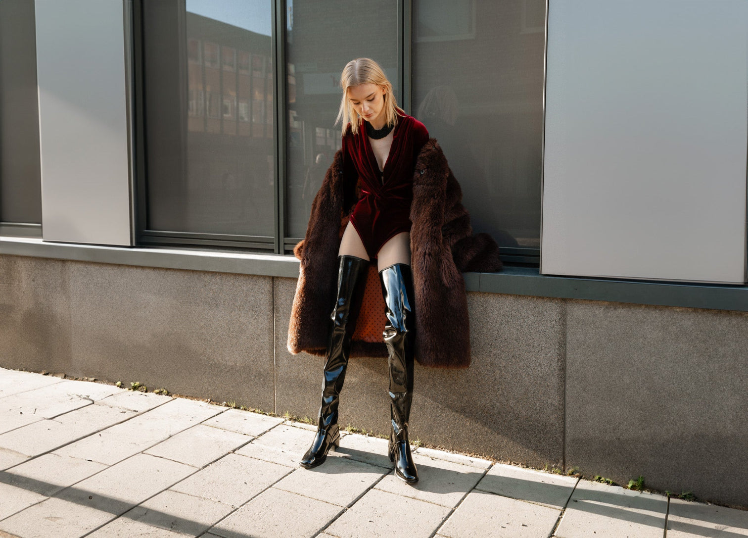 Fashion-forward model wearing ACQUO of Sweden over-the-knee glossy black rubber boots styled with a deep red velvet bodysuit and luxurious coat, showcasing a bold and elegant street style look perfect for modern, confident women.