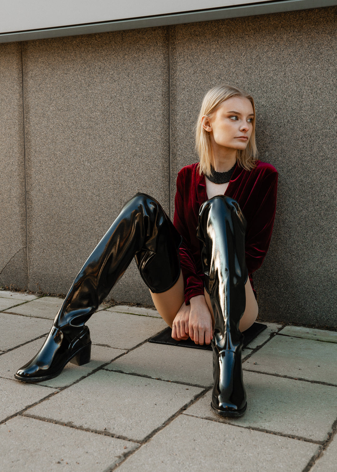 Model wearing ACQUO of Sweden over-the-knee glowing black heeled boots, styled with a deep red velvet bodysuit and black statement necklace – a bold and elegant street style look with luxurious texture contrast