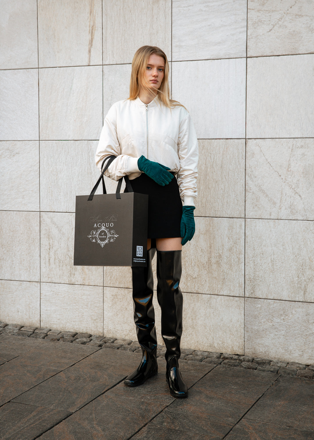 Model wearing ACQUO of Sweden over-the-knee glowing black boots, styled with a cream bomber jacket, black mini skirt, teal gloves, and holding a branded ACQUO shopping bag – a modern and elegant street style look with luxury fashion flair