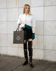 Model wearing ACQUO of Sweden over-the-knee glowing black boots, styled with a cream bomber jacket, black mini skirt, teal gloves, and holding a branded ACQUO shopping bag – a modern and elegant street style look with luxury fashion flair