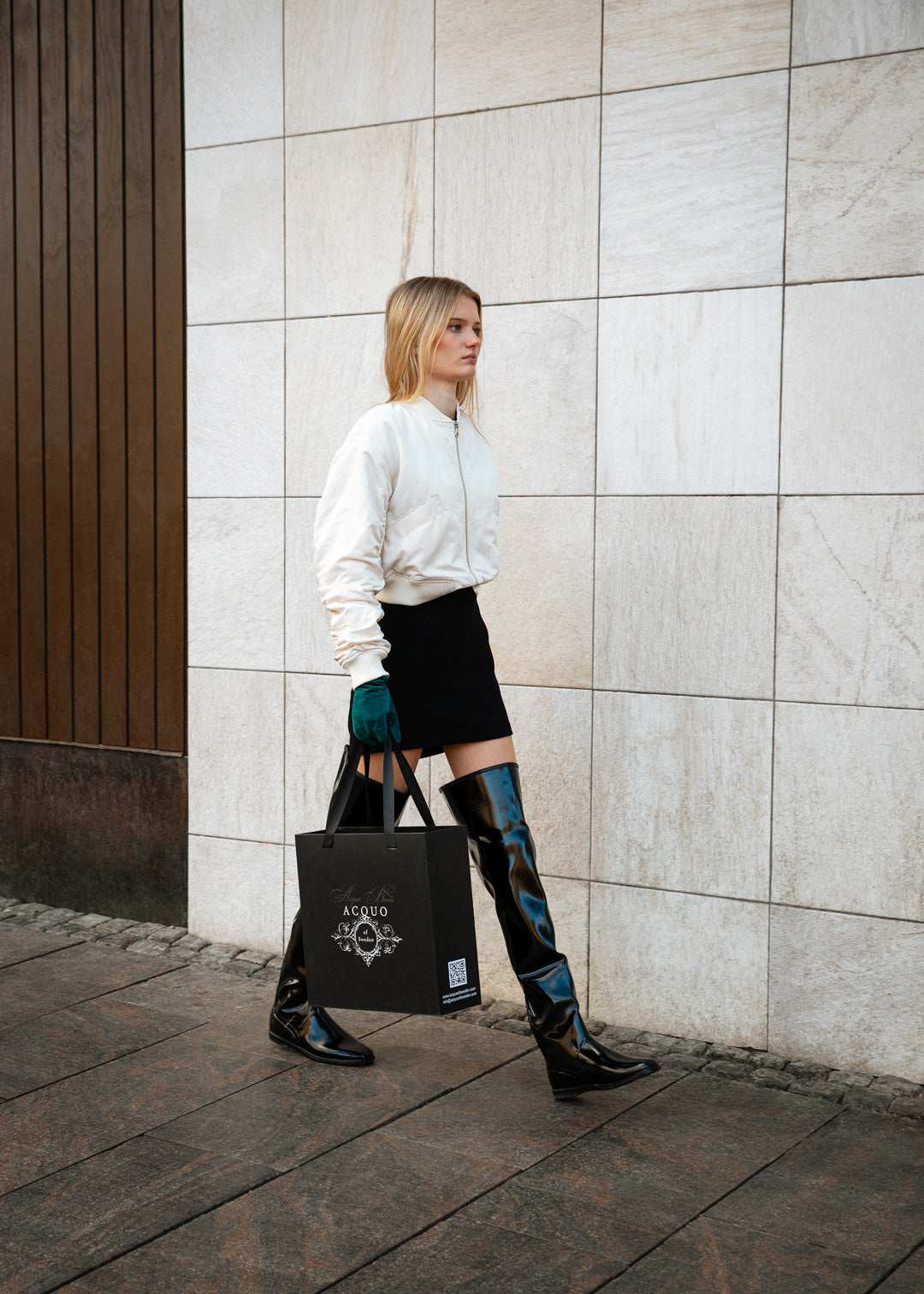Stylish woman walking confidently along a stone wall in a chic urban outfit, wearing black over-the-knee ACQUO of Sweden boots, a white bomber jacket, and a black mini skirt, while holding an elegant black shopping bag with the ACQUO logo—embodying modern Swedish fashion with timeless elegance