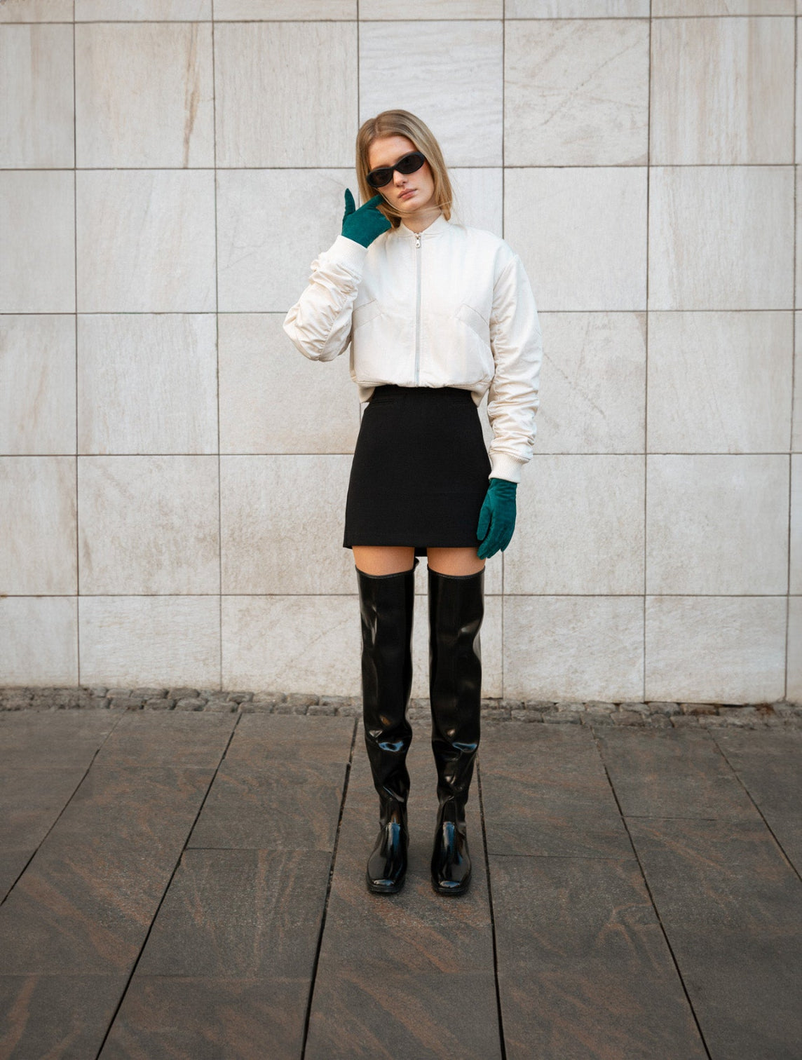 Fashion-forward woman in a monochrome outfit featuring black over-the-knee ACQUO of Sweden boots, a black mini skirt, white bomber jacket, dark sunglasses, and green gloves, posing confidently against a minimalist stone wall—highlighting modern Scandinavian style with bold, elegant footwear