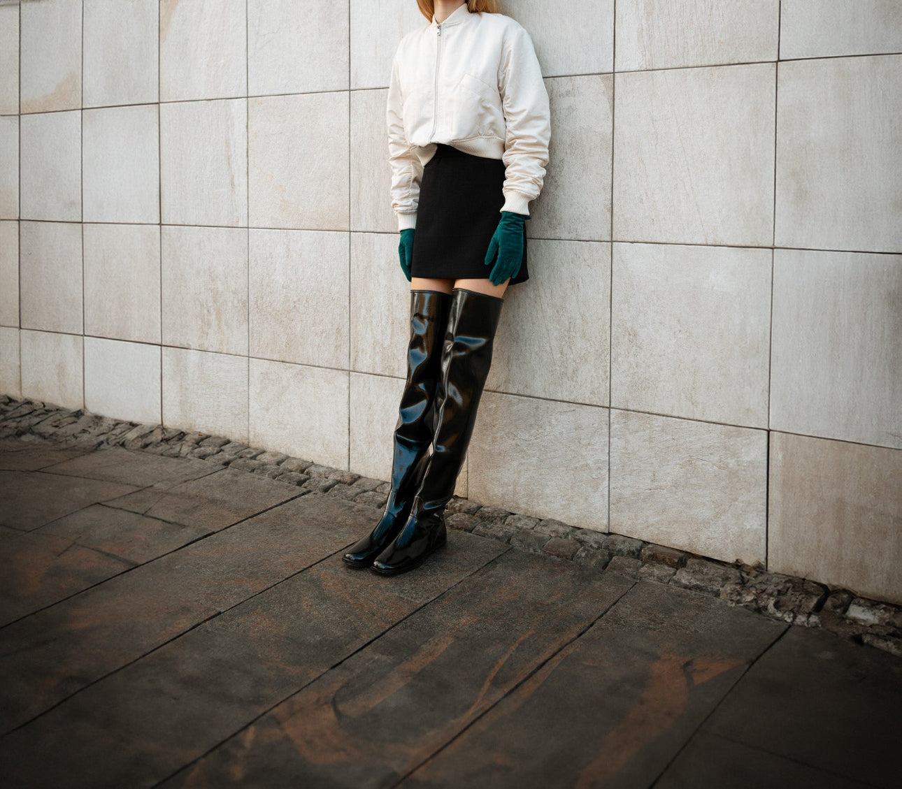 Close-up of ACQUO of Sweden's signature glowing black over-the-knee boots styled with a black mini skirt, ivory bomber jacket, and emerald green gloves—modern Scandinavian fashion combining elegance and attitude in high-shine rubber footwear