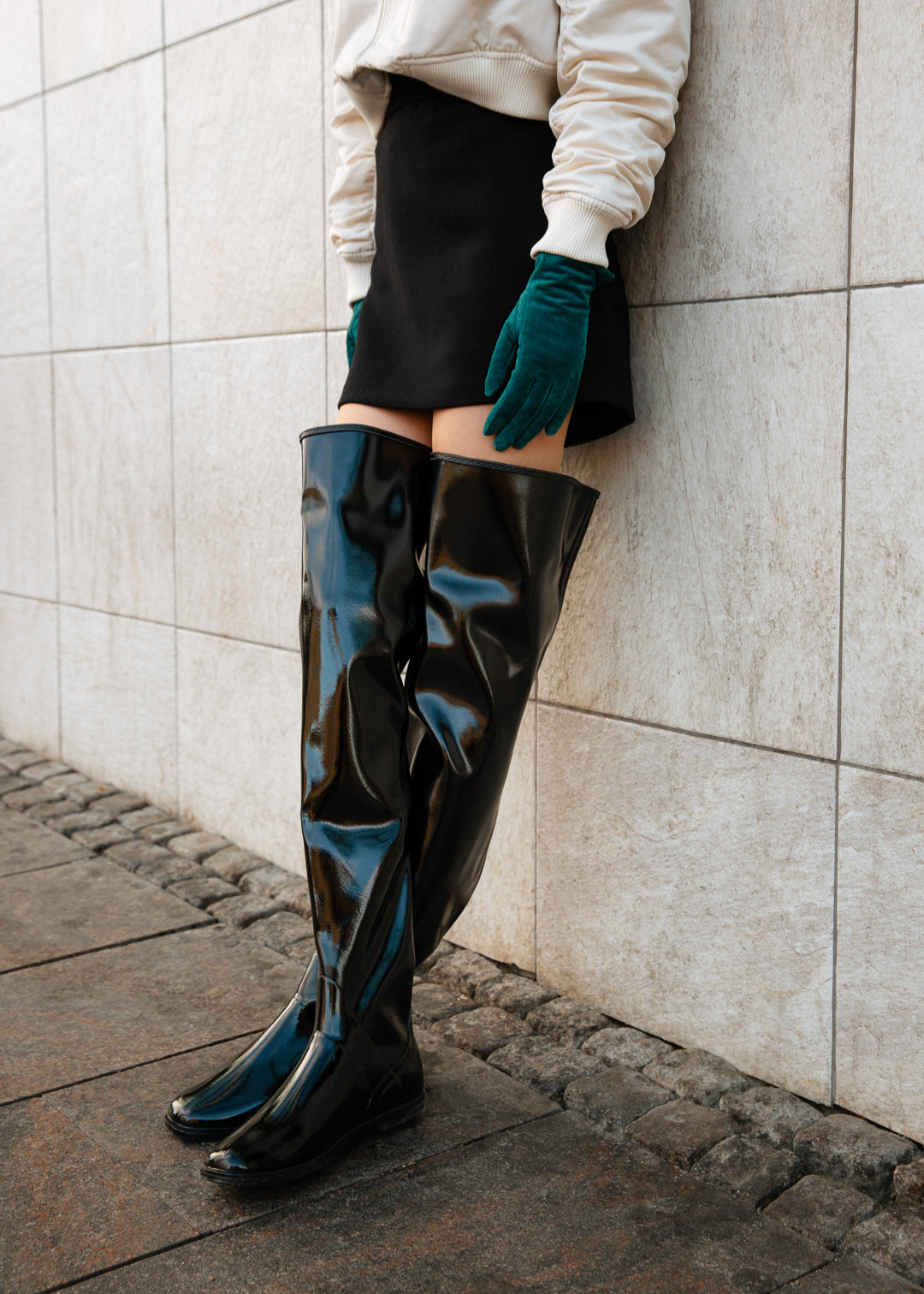 Close-up of model wearing ACQUO of Sweden over-the-knee glowing black boots with a flat sole, styled with a black mini skirt, cream bomber jacket, and teal suede gloves – a bold and polished urban fashion statement