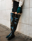 Close-up of model wearing ACQUO of Sweden over-the-knee glowing black boots with a flat sole, styled with a black mini skirt, cream bomber jacket, and teal suede gloves – a bold and polished urban fashion statement