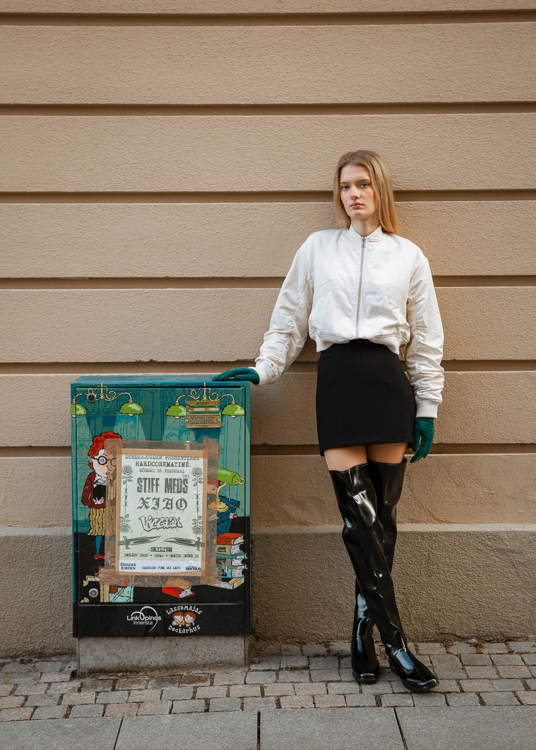 Model wearing ACQUO of Sweden over-the-knee glowing black boots, paired with a black mini skirt, cream bomber jacket, and teal gloves – striking urban fashion captured against a beige wall and colorful street art panel