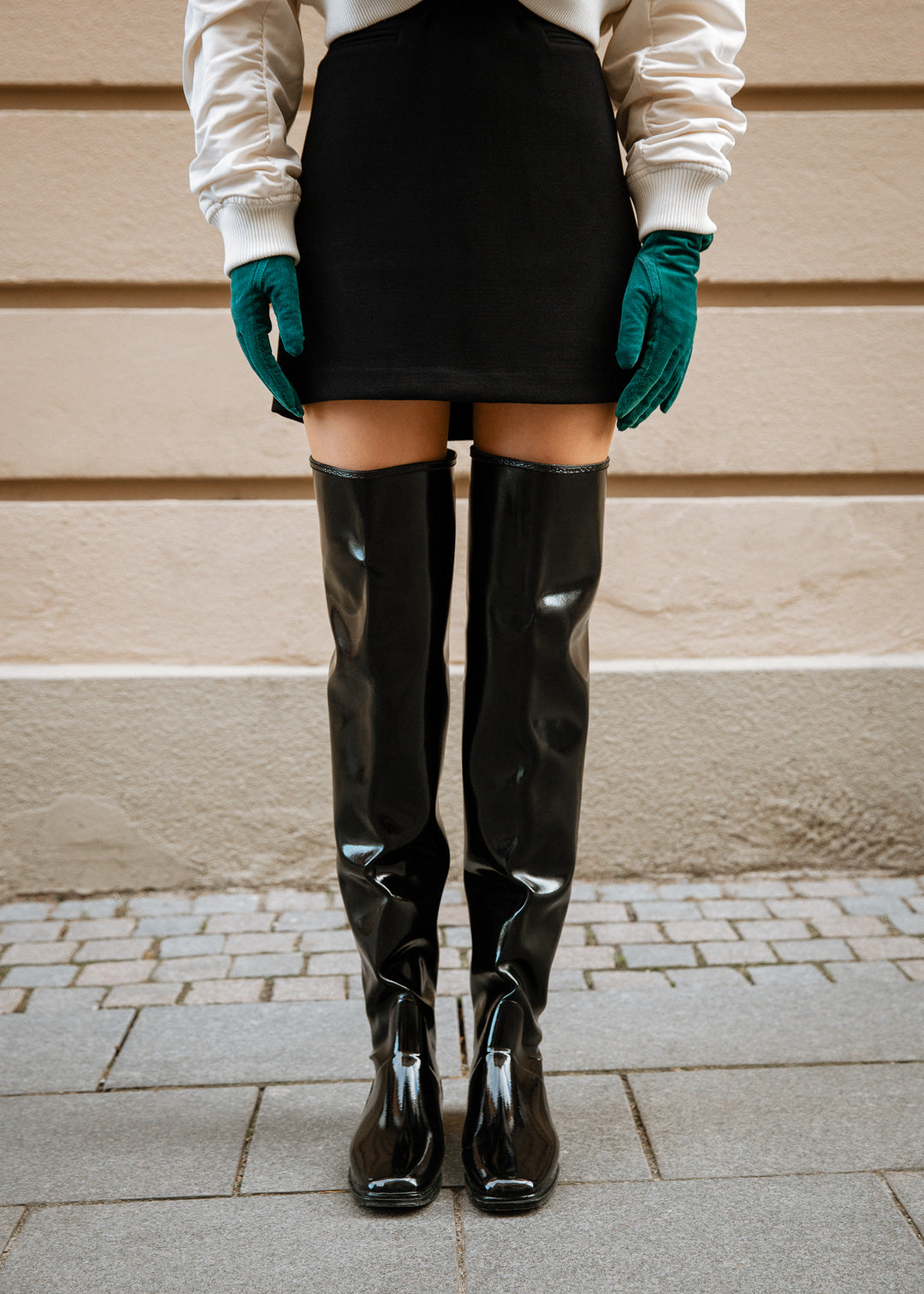 Woman wearing black over-the-knee rubber boots from ACQUO of Sweden, styled with a black mini skirt, white bomber jacket, and teal gloves—capturing a modern Scandinavian fashion look in an urban setting
