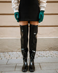 Woman wearing black over-the-knee rubber boots from ACQUO of Sweden, styled with a black mini skirt, white bomber jacket, and teal gloves—capturing a modern Scandinavian fashion look in an urban setting
