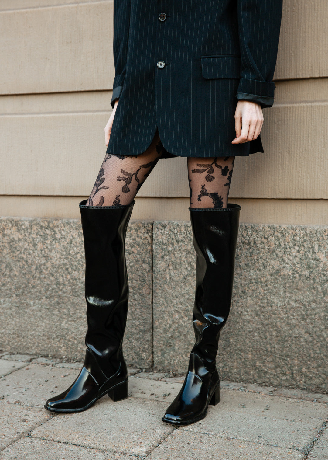 Close-up of a stylish outfit featuring ACQUO of Sweden’s signature black over the knee rubber boots, paired with sheer floral patterned tights and a pinstripe blazer dress—capturing contemporary Scandinavian fashion with a bold and elegant street style edge