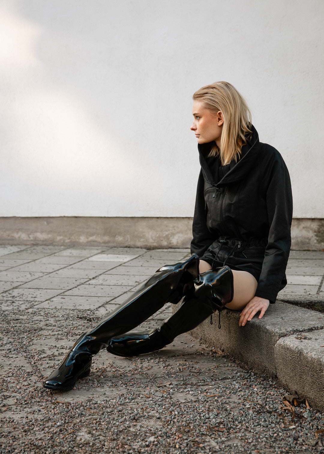 Model wearing ACQUO of Sweden over-the-knee glowing black boots with a flat sole, styled with a black jacket and shorts – a bold, minimalist street style look in an urban outdoor setting