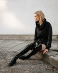 Model wearing ACQUO of Sweden over-the-knee glowing black boots with a flat sole, styled with a black jacket and shorts – a bold, minimalist street style look in an urban outdoor setting