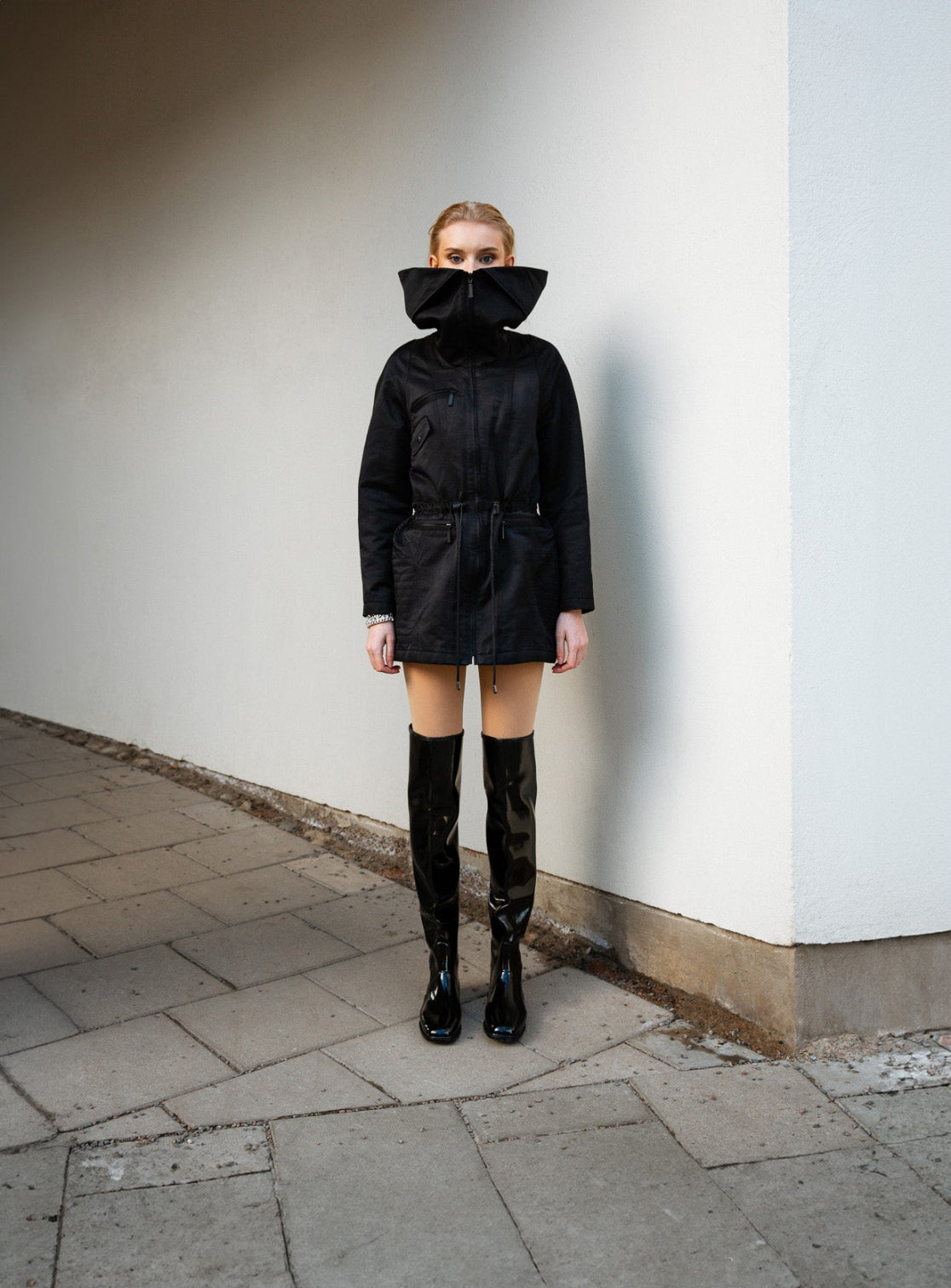 A fashion-forward woman stands confidently in a black high-collar jacket and over the knee black boots by ACQUO of Sweden, showcasing a bold Scandinavian style statement. The image highlights ACQUO’s signature high-shine rubber boots in an urban minimalist setting
