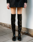 Close-up of a model wearing glowing black over-the-knee boots by ACQUO of Sweden, paired with a short black coat—elegant Swedish design in high-shine natural rubber for bold and stylish statements
