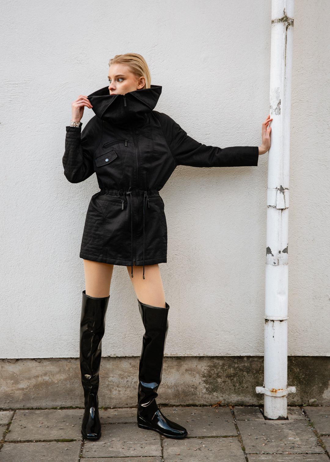 Model wearing ACQUO of Sweden over-the-knee glowing black boots with a flat sole, paired with a short black utility-style jacket – a bold, modern look set against a minimalist urban background