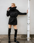 Model wearing ACQUO of Sweden over-the-knee glowing black boots with a flat sole, paired with a short black utility-style jacket – a bold, modern look set against a minimalist urban background