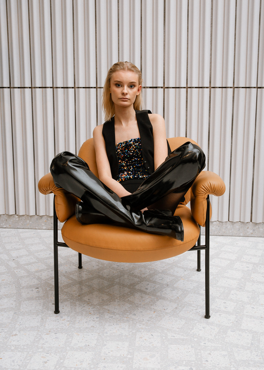 Woman seated cross-legged in a modern tan armchair wearing black over-the-knee ACQUO of Sweden boots paired with a sparkly top and vest—showcasing contemporary Swedish fashion design with bold elegance and distinctive style
