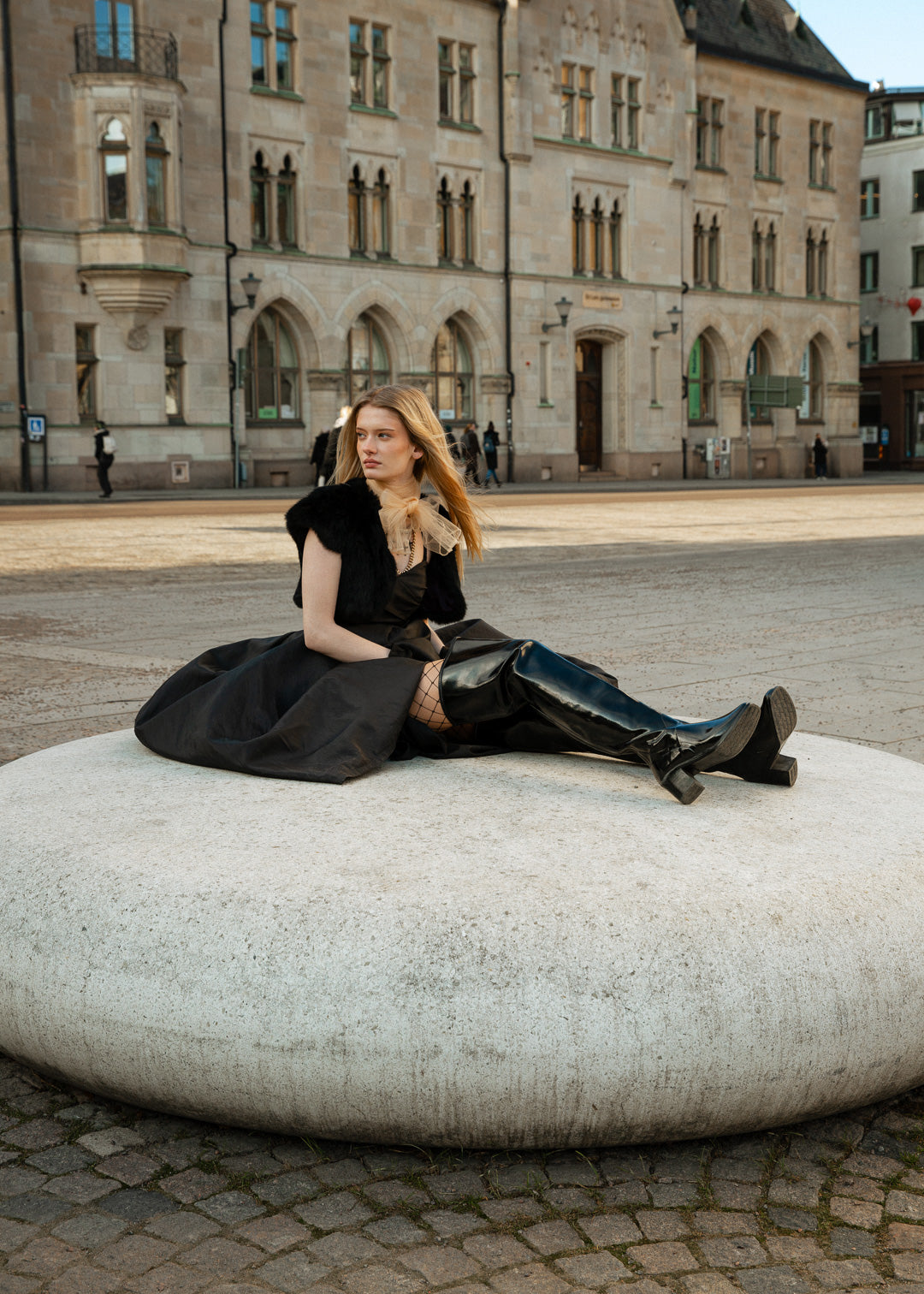 Fashionable woman sitting on a modern stone sculpture in an urban square, wearing black over-the-knee ACQUO of Sweden boots paired with a voluminous black skirt, fishnet tights, and a soft beige bow scarf—an elegant fusion of classic and contemporary Swedish style