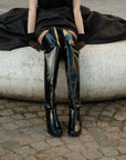 Model sitting on a sculptural stone bench wearing ACQUO of Sweden over-the-knee black rubber boots with a statement flare, paired with a dramatic black skirt and sheer gloves—luxurious Scandinavian footwear blending elegance and attitude