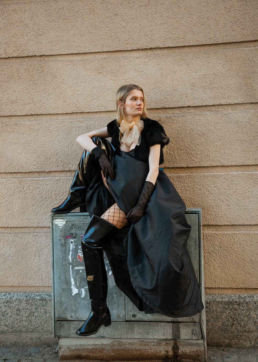 Model wearing ACQUO of Sweden over-the-knee glowing black boots with heels, styled with a dramatic black gown, fishnet stockings, sheer gloves, and a beige tulle neck bow – a bold blend of high fashion and urban edge in a Scandinavian city setting