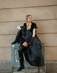 Model wearing ACQUO of Sweden over-the-knee glowing black boots with heels, styled with a dramatic black gown, fishnet stockings, sheer gloves, and a beige tulle neck bow – a bold blend of high fashion and urban edge in a Scandinavian city setting