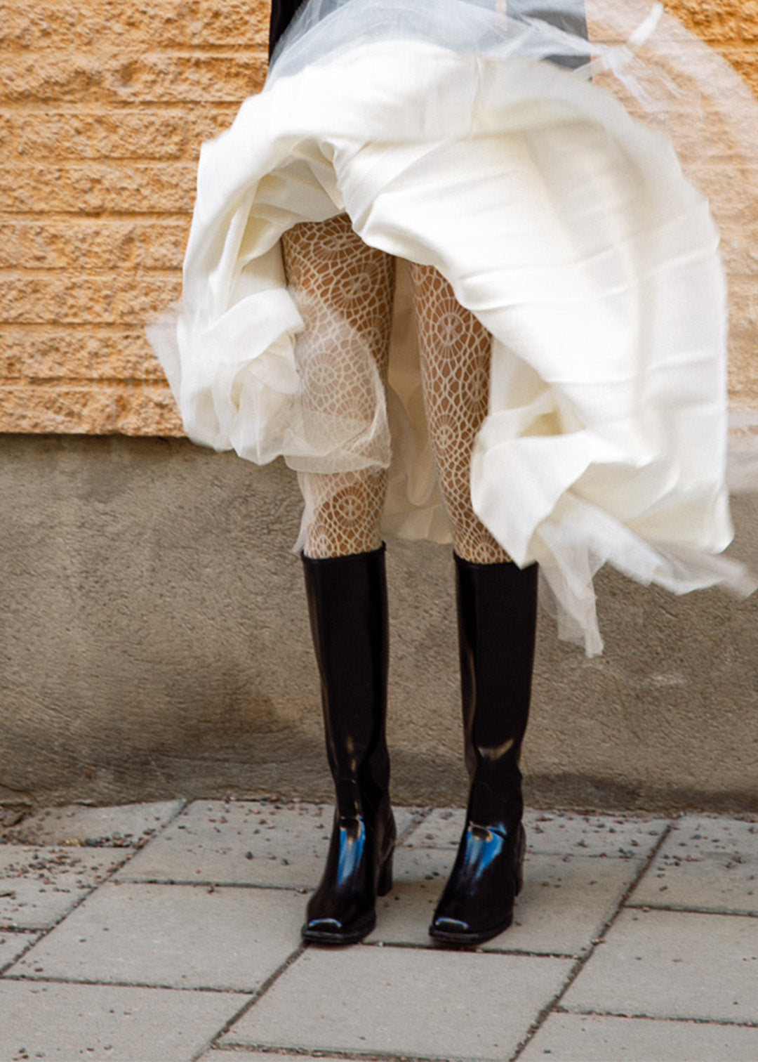 Woman wearing knee-high black rubber boots from ACQUO of Sweden paired with patterned white tights and a flowing layered white skirt, captured in motion against a rustic stone wall—an elegant fusion of vintage and modern Scandinavian style