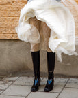 Woman wearing knee-high black rubber boots from ACQUO of Sweden paired with patterned white tights and a flowing layered white skirt, captured in motion against a rustic stone wall—an elegant fusion of vintage and modern Scandinavian style