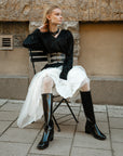 Model in ACQUO of Sweden's knee-high sleek shine black boots with a block heel, paired with a flowing white tulle skirt, textured white tights, and a sleek black blouse – blending feminine elegance with edgy street style