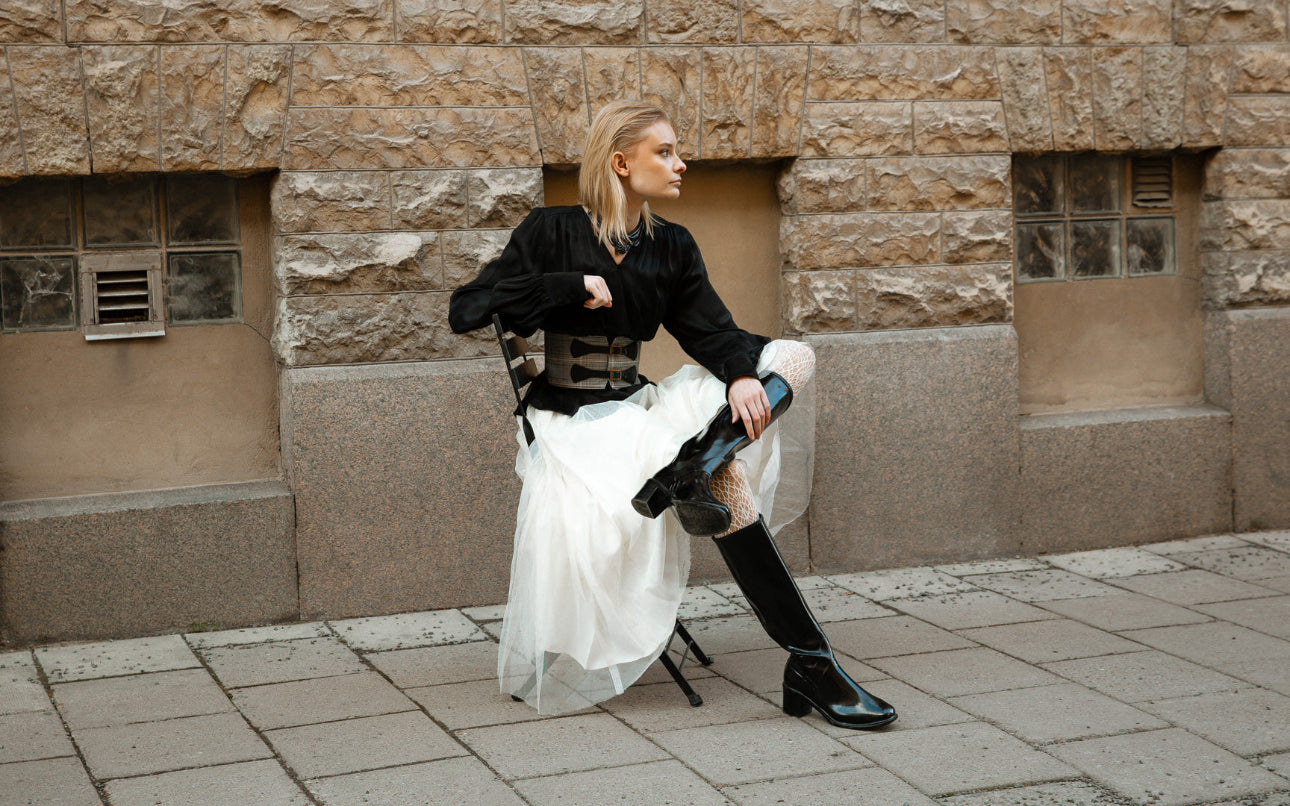 Stylish woman in ACQUO of Sweden's signature black the knee high rubber boots paired with a flowing white tulle skirt and structured black blouse, sitting confidently on a city street against a stone wall, showcasing bold Swedish fashion and premium boot design.