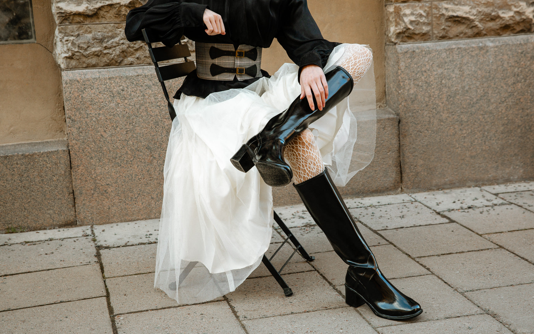 Model wearing ACQUO of Sweden high-shine black boots with block heels, styled with a voluminous white tulle skirt, fishnet tights, and a wide statement belt – a bold fusion of elegance and Scandinavian fashion edge