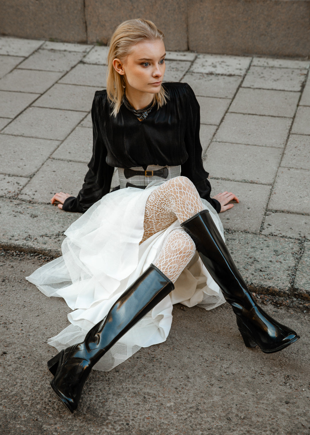 Model wearing ACQUO of Sweden knee-high glowing black boots with block heel, styled with a white layered tulle skirt, patterned white tights, and a black long-sleeve top – a bold and elegant street style look