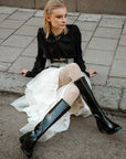 Model wearing ACQUO of Sweden knee-high glowing black boots with block heel, styled with a white layered tulle skirt, patterned white tights, and a black long-sleeve top – a bold and elegant street style look