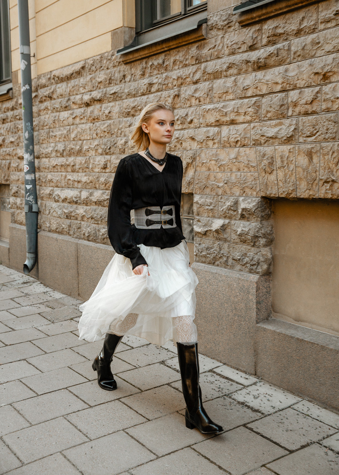 Model showcases ACQUO of Sweden knee-high glowing black boots with a block heel, styled with a voluminous white tulle skirt, lace-patterned tights, and a structured black blouse – a striking fusion of modern elegance and streetwear attitude