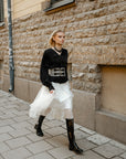 Model showcases ACQUO of Sweden knee-high glowing black boots with a block heel, styled with a voluminous white tulle skirt, lace-patterned tights, and a structured black blouse – a striking fusion of modern elegance and streetwear attitude