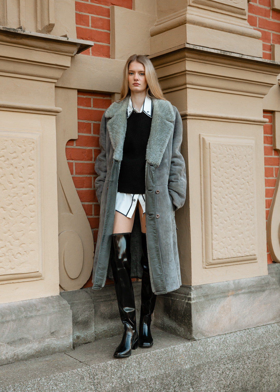 Model wearing ACQUO of Sweden over-the-knee glowing black boots, styled with a white shirt dress, black knit vest, and long grey shearling coat – a chic layered look combining modern fashion with classic Scandinavian outerwear