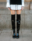 Close-up of model wearing ACQUO of Sweden over-the-knee glowing black boots, styled with a white shirt dress and black knitted vest – modern, statement footwear with a Scandinavian street style edge