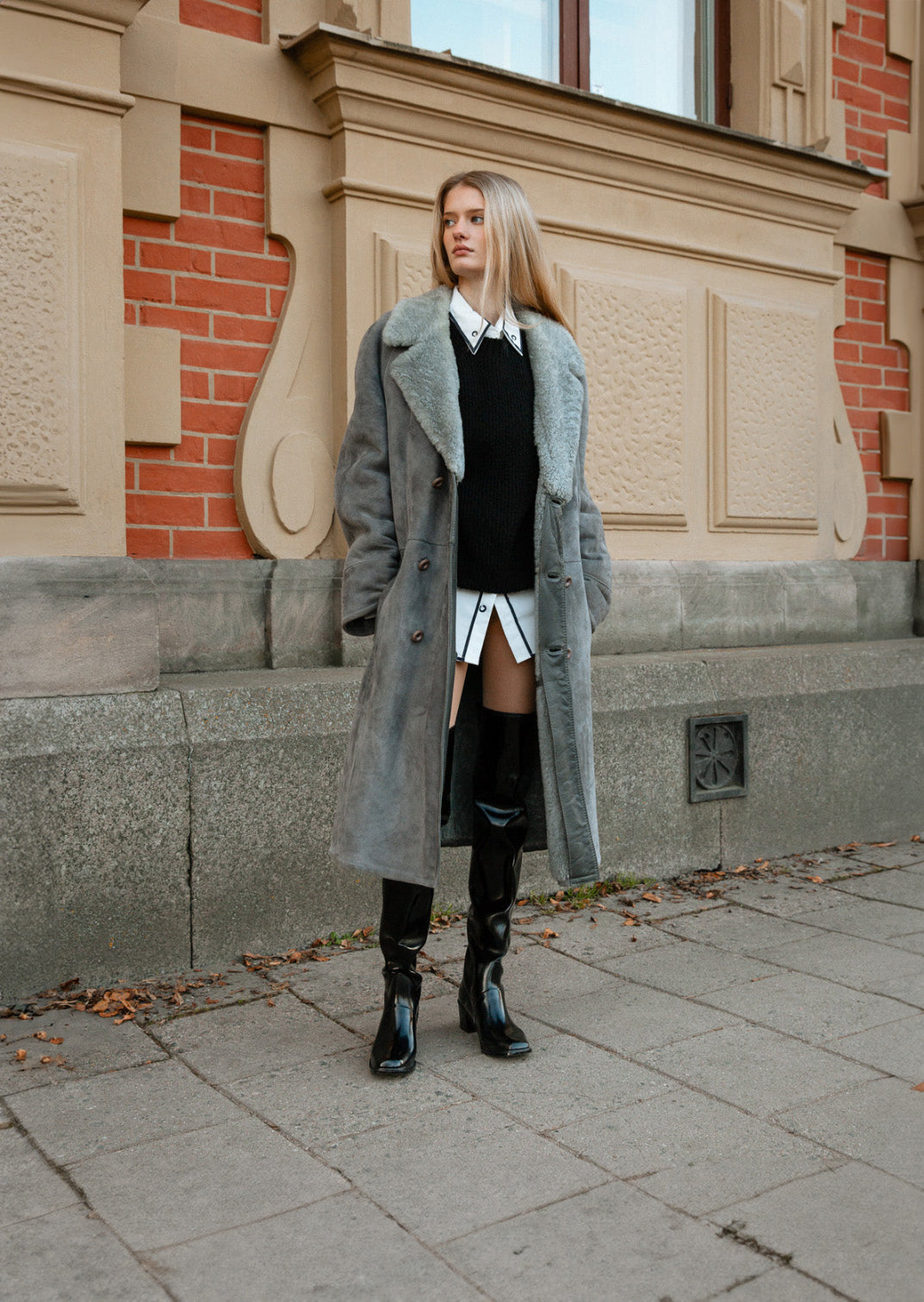 Fashionable woman standing in front of a historic building wearing ACQUO of Sweden over the knee black boots, a long grey shearling coat, and a layered black and white outfit — a sophisticated autumn street style look that highlights the bold, elegant design of ACQUO boots
