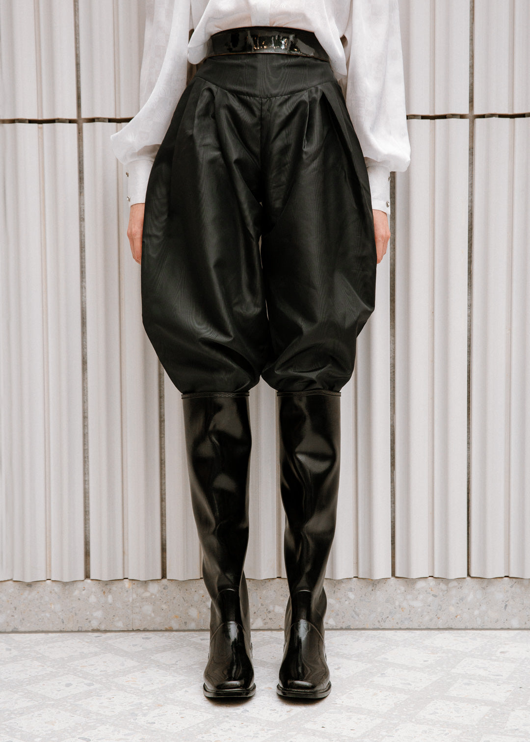 Close-up of a fashion-forward outfit featuring ACQUO of Sweden’s signature black the knee high rubber boots, styled with voluminous black trousers and a wide patent belt, paired with a crisp white blouse—set against a minimalist indoor backdrop with vertical paneling