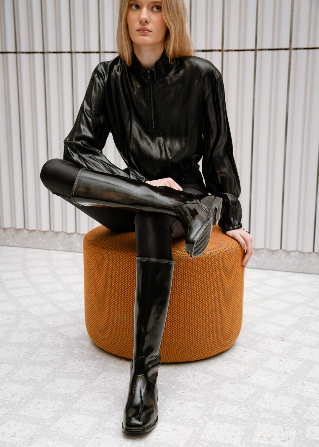 Model wearing ACQUO of Sweden glossy black boots, paired with a black patent-look top and tights, sitting on an orange designer stool