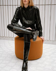 Model wearing ACQUO of Sweden glossy black boots, paired with a black patent-look top and tights, sitting on an orange designer stool