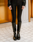 Front view of ACQUO of Sweden's sleek knee-high black rubber boots styled with fitted black trousers and a satin blouse—timeless Scandinavian craftsmanship meets contemporary fashion