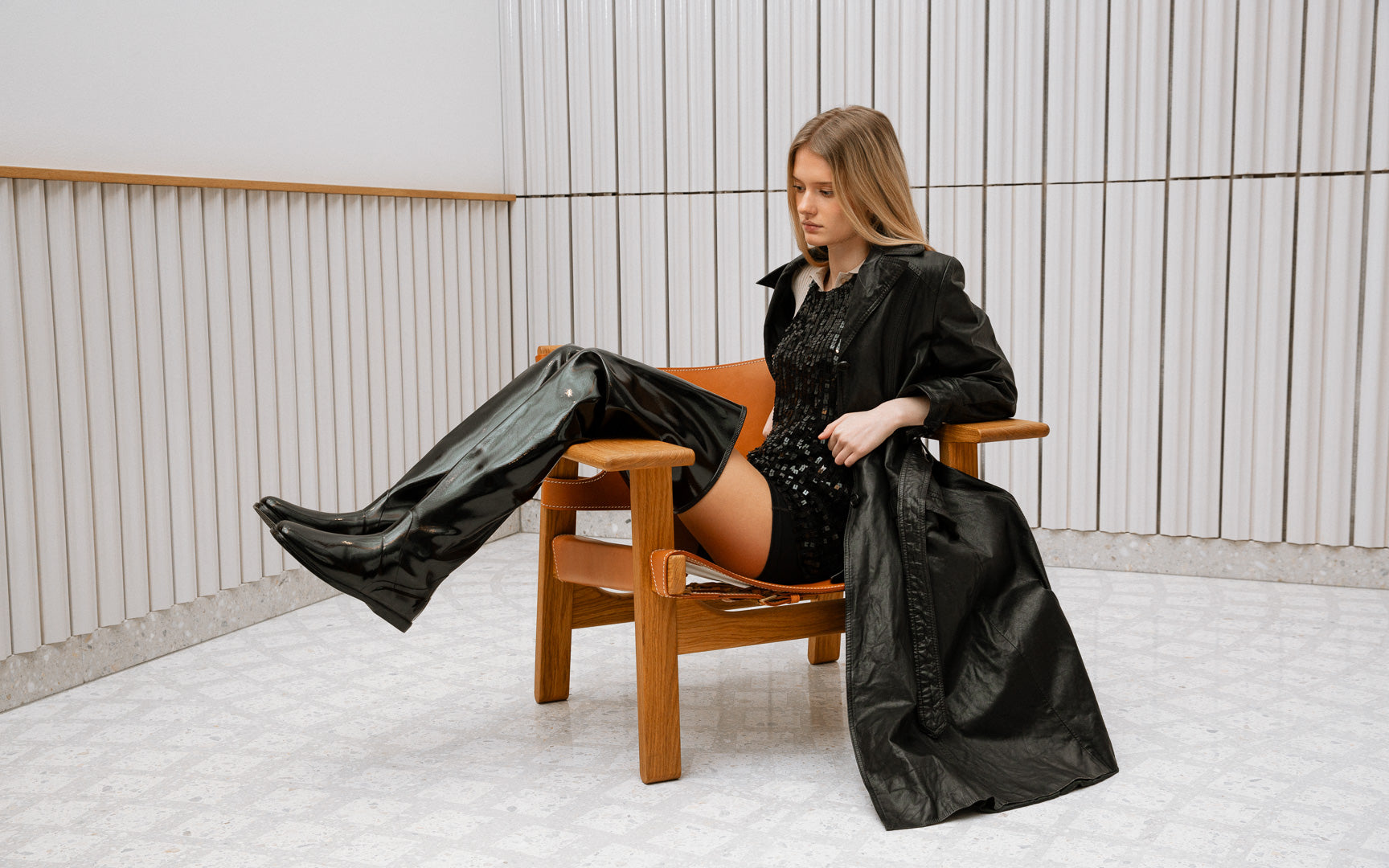 Model wearing ACQUO of Sweden over-the-knee black boots paired with a black sequin top and long trench coat, seated in a Scandinavian modern chair – bold, elegant fashion with a retro-luxury edge