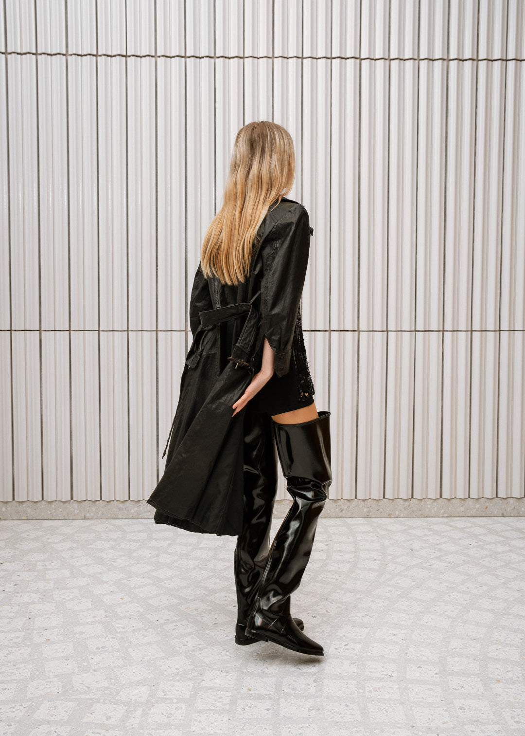 Model dressed in ACQUO of Sweden high-shine over-the-knee black boots, paired with a black embellished top, short bottoms, and a flowing black trench coat – a bold and elegant look set in a modern Scandinavian space