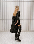 Model dressed in ACQUO of Sweden high-shine over-the-knee black boots, paired with a black embellished top, short bottoms, and a flowing black trench coat – a bold and elegant look set in a modern Scandinavian space