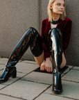 Model wearing ACQUO of Sweden over-the-knee glowing black heeled boots, styled with a deep red velvet bodysuit and black statement necklace – a bold and elegant street style look with luxurious texture contrast
