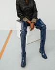 Model seated on white display cubes wearing Elsa Iris Blue boots by ACQUO of Sweden. The image shows tall iris blue rubber boots with a sleek fitted silhouette, square toe, and low block heel, styled with a dark patterned jacket in a minimalist studio setting