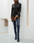Model leaning against a white wall wearing Elsa Iris Blue boots by ACQUO of Sweden. The image shows elegant iris blue over-the-knee rubber boots with a sleek fitted silhouette, square toe, and reflective finish, styled with patterned tights and a tailored black jacquard jacket in a minimalist studio interior.
