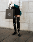 Model wearing ACQUO of Sweden over-the-knee glowing black boots, styled with a cream bomber jacket, black mini skirt, teal gloves, and holding a branded ACQUO shopping bag – a modern and elegant street style look with luxury fashion flair